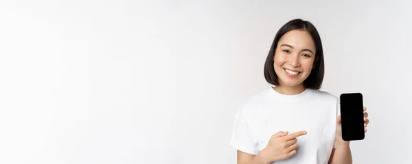 Usmívající se asijská žena ukazuje prst na obrazovce smartphone, ukazuje aplikační rozhraní, mobilní telefon webové stránky, stojící nad bílým pozadím — Stock fotografie