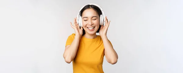 행복 한 동양인 소녀 춤, 헤드폰으로 음악을 듣고 웃는 모습, 노란 티셔츠를 입고 하얀 배경을 배경으로 서 있는 모습 — 스톡 사진