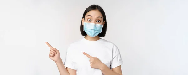Förvånad asiatisk flicka i medicinsk mask, pekande fingrar vänster, visar promo erbjudande, höja ögonbryn, förvånad reaktion, står mot vit bakgrund — Stockfoto