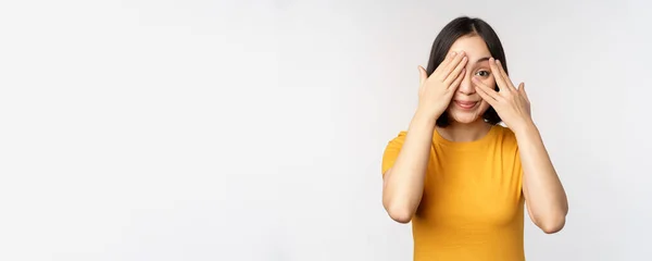 Söt koreansk kvinnlig modell, täck ögonen med händerna, kika genom fingrarna nyfiken, titta på smth, står i gul t-shirt över vit bakgrund — Stockfoto