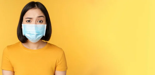 Retrato de cético e confuso ásia mulher no médico rosto máscara levantando sobrancelha duvidoso de pé sobre amarelo fundo — Fotografia de Stock
