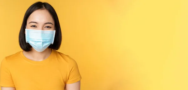 Blisko uśmiechnięta szczęśliwa Azjatka, ubrana w medyczną maskę na twarz od covid-19, stojąca w żółtej koszulce na tle studia — Zdjęcie stockowe