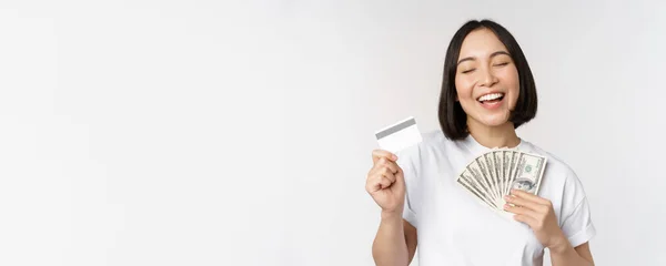 Portrait of asian woman smiling, holding credit card and money cash, dollars, standing in tshirt over white background — Stock Photo, Image