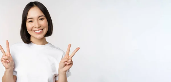 Tatlı Asyalı kız barış, V-işareti, gülümsüyor ve kameraya mutlu bakıyor, beyaz tişört giyiyor, stüdyo arka planı var. — Stok fotoğraf