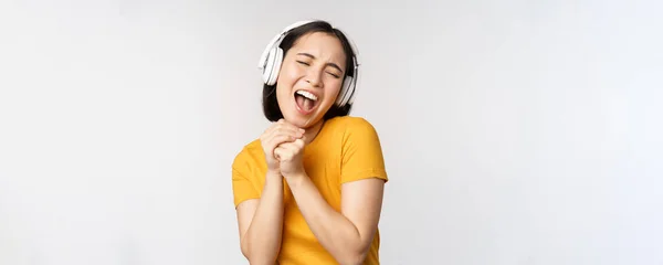 Felice ragazza asiatica che balla, ascolta musica su cuffie e sorride, in piedi in maglietta gialla sullo sfondo bianco — Foto Stock