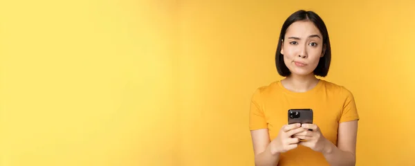 Skeptisk asiatisk kvinna som håller smartphone, ser med tvivel på kameran, står över gul bakgrund — Stockfoto