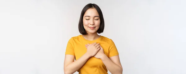 Novia asiática romántica, cogida de la mano en el corazón, sonriendo con cuidado y ternura, de pie en camiseta amarilla sobre fondo blanco — Foto de Stock