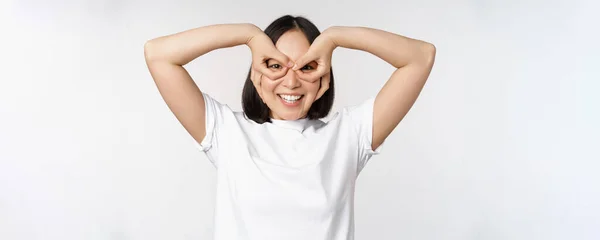 Vtipný mladý asijské žena, korejština dívka dělat oči brýle gesto, pohled šťastný na kameru, stojící nad bílým pozadím — Stock fotografie