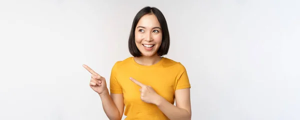 Portrait of smiling asian brunette girl in yellow tshirt, pointing fingers left, showing copy space, promo deal, demonstrating banner, standing over white background — Stock Photo, Image