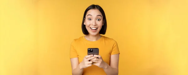 Felice ragazza asiatica sorridente, in piedi con il telefono cellulare nero, in piedi contro sfondo giallo — Foto Stock