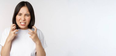 Kızgın Asyalı kadın küfür ediyor, öfkeli ve sinirli görünüyor, dişlerini sıkmış ve kaşlarını çatmış, beyaz arka planda duruyor.