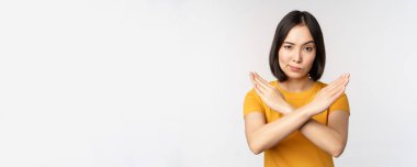Asyalı kadın portresi ciddi ve sinirli görünüyor, dur işareti, tabu işareti, müstehcenliği yasaklıyor, beyaz arka plan üzerinde sarı tişörtle duruyor.