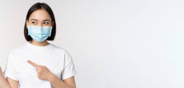 Tıbbi maske takan genç Koreli kadın solu işaret ediyor ve logoya bakıyor, reklam ya da afiş gösteriyor, beyaz arka planda duruyor.