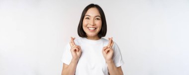 Umut dolu Asyalı kız parmaklarını çapraz yapıyor, dilek tutuyor, umut ediyor, pürüzsüz olmasını diliyor, beyaz arka planda tişörtün üzerinde duruyor.