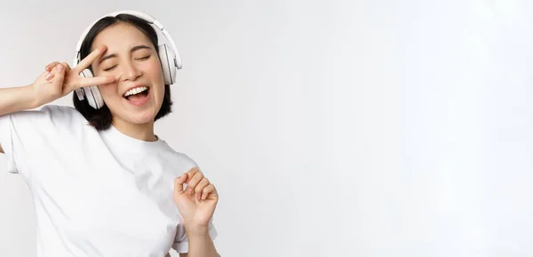 Bailando y cantando mujer asiática, escuchando música en auriculares, de pie en auriculares sobre fondo blanco — Foto de Stock