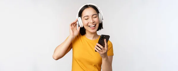 Menina japonesa bonito em fones de ouvido, olhando para o telefone celular e sorrindo, usando aplicativo de música no smartphone, de pé contra fundo branco — Fotografia de Stock