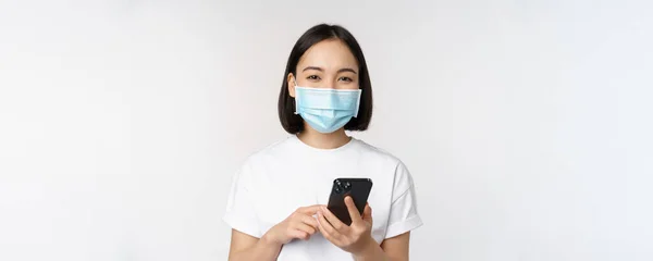 Saúde e conceito covid-19. Menina asiática em máscara facial médica, usando o aplicativo de telefone móvel em quarentena, de pé sobre fundo branco — Fotografia de Stock