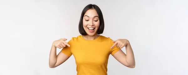 Feliz chica asiática señalando con los dedos hacia abajo, mirando anuncio, pancarta o anuncio, de pie en camiseta amarilla sobre fondo blanco —  Fotos de Stock