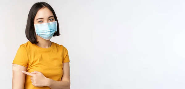 Covid-19, aşı ve sağlık konsepti. Tıbbi maskeli sevimli Asyalı kızın portresi, beyaz arka planda duran, Coronavirus aşısından sonra omuzlarında yara bandı var. — Stok fotoğraf