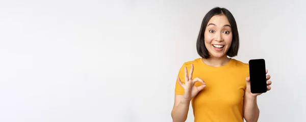 Aufgeregte Asiatin zeigt Handybildschirm, Okay-Schild, empfiehlt Smartphone-App, steht in gelbem T-Shirt vor weißem Hintergrund — Stockfoto