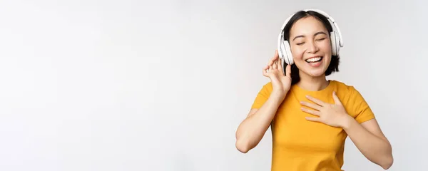 Szczęśliwa Azjatka tańczy, słucha muzyki na słuchawkach i uśmiecha się, stojąc w żółtej koszuli na białym tle — Zdjęcie stockowe