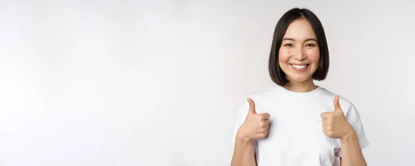 Šťastná mladá korejská žena ukazuje palce nahoru a usmívá se, dát pozitivní zpětnou vazbu, doporučuje smth, stojící nad bílým pozadím — Stock fotografie