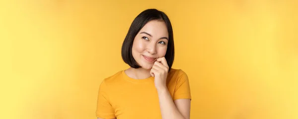 Κοντινό πλάνο πορτρέτο του χαριτωμένο κορίτσι της Ασίας χαμογελώντας, σκέψης, κοιτάζοντας μέχρι στοχαστική, στέκεται σε μπλουζάκι πάνω από κίτρινο φόντο — Φωτογραφία Αρχείου