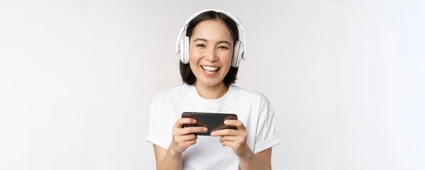 Glad asiatisk flicka gamer, spela på mobiltelefon, titta på smartphone, bär hörlurar, står över vit bakgrund — Stockfoto