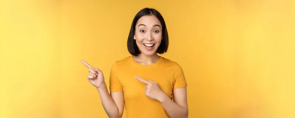 Mujer asiática sonriente señalando con los dedos a la izquierda, mostrando publicidad en espacio de copia vacío, de pie sobre fondo amarillo — Foto de Stock