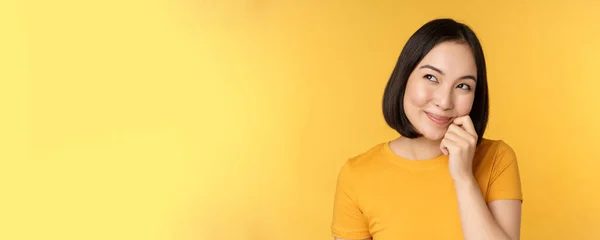 Primer plano retrato de linda chica asiática sonriendo, pensando, mirando hacia arriba reflexivo, de pie en camiseta sobre fondo amarillo — Foto de Stock