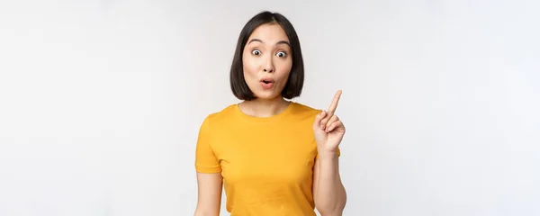 Schöne junge Asiatin zeigt mit dem Finger nach oben, lächelt und schaut amüsiert in die Kamera, zeigt Werbung, oben Ankündigung, weißer Hintergrund — Stockfoto