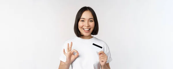 Chica coreana sonriente mostrando signo de aprobación y recomendando tarjeta de crédito del banco de espacio de copia, de pie en camiseta sobre fondo blanco — Foto de Stock