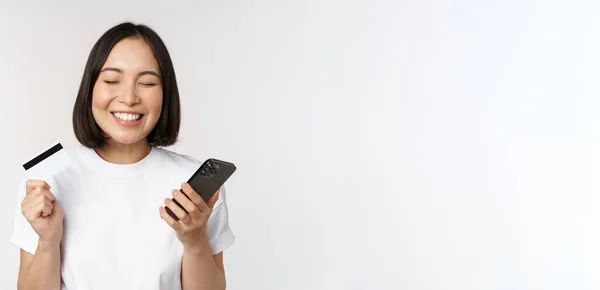 Online shopping. Happy asian woman using credit card and smartphone app, paying on website via mobile phone, white background — Stock Photo, Image