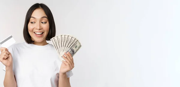 Portrait of asian woman smiling, holding credit card and money cash, dollars, standing in tshirt over white background — Stock Photo, Image