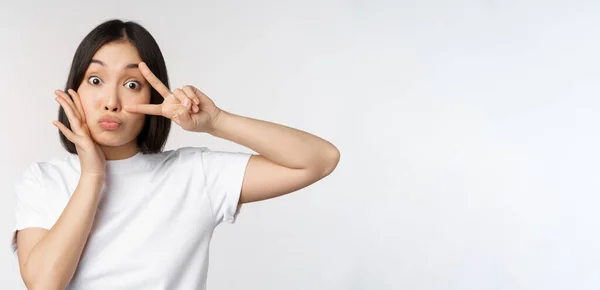 Cute asian girl posing with kawaii v-sign, peace gesture near face, standing in tshirt over white background — Stock Photo, Image