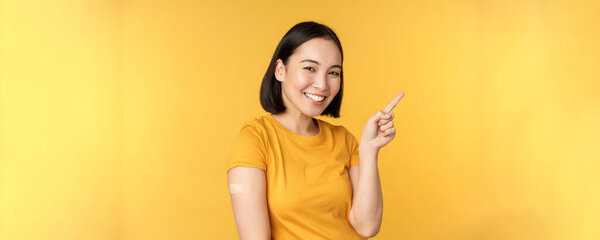 Vaccination and covid-19 pandemic concept. Smiling korean woman with band aid on shoulder after coronavirus vaccine shot, pointing at banner with vaccinating campaign