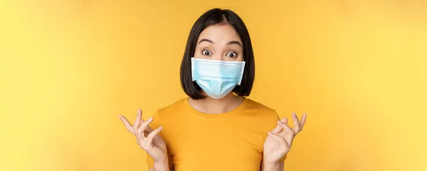 Covid-19 and medical concept. Close up portrait of asian woman in face mask, looking surprised and amazed at news, standing over yellow background — Stock Photo, Image