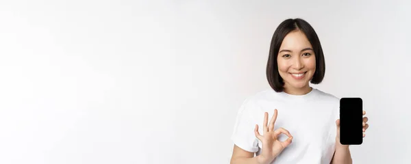 Lächelnde Asiatin zeigt Okay, Handybildschirm, empfiehlt Smartphone-App, steht über weißem Hintergrund — Stockfoto