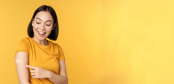 Вакцинация и концепция пандемии ковида-19. Счастливая и здоровая азиатская девушка указывая на ее плечо с помощью браслета после вакцинации от коронавируса, желтый фон — стоковое фото