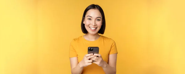 Tecnología. Mujer asiática sonriente usando teléfono móvil, sosteniendo el teléfono inteligente en las manos, de pie en camiseta sobre fondo amarillo — Foto de Stock