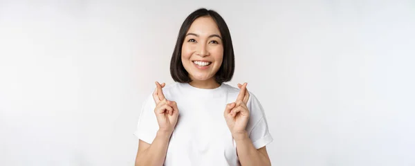 Esperanzado chica asiática cruzar los dedos, pidiendo deseo, anticipando, deseando smth, de pie en camiseta sobre fondo blanco — Foto de Stock