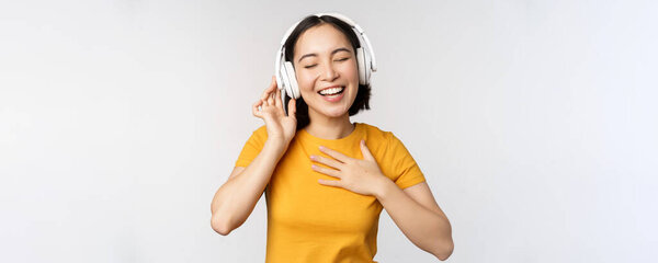 Счастливая азиатская девушка танцует, слушает музыку на наушниках и улыбается, стоя в желтой футболке на белом фоне