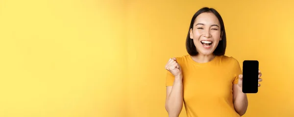 Spännande asiatisk kvinna, visar smartphone app och triumferande, firar på mobiltelefon, står över gul bakgrund — Stockfoto