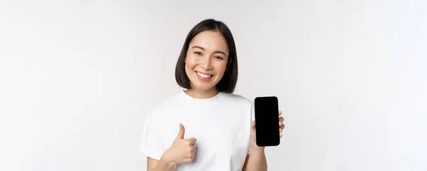 Portret van een mooie Aziatische vrouw met duim omhoog en smartphone scherm, glimlachen tijdens het adverteren mobiele telefoon app, witte achtergrond — Stockfoto