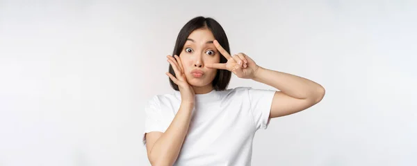 Aranyos ázsiai lány pózol Kawaii v-jel, béke gesztus közel arc, álló pólóban fehér háttér — Stock Fotó