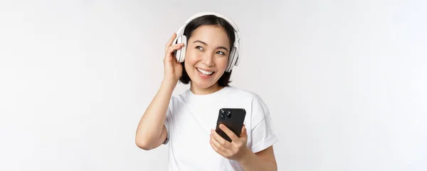 Hermosa chica asiática moderna, escuchando música en auriculares, sosteniendo el teléfono móvil, utilizando la aplicación para teléfonos inteligentes, de pie sobre fondo blanco — Foto de Stock