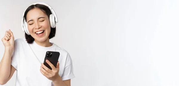Schöne moderne asiatische Mädchen, Musik über Kopfhörer hören, Handy in der Hand, Smartphone-App, vor weißem Hintergrund stehen — Stockfoto