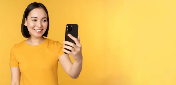 Bild av glad, vacker asiatisk flicka videochatt, talar på smartphone ansökan, står mot gul bakgrund — Stockfoto