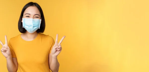 Θετική Ασιάτισσα χαμογελαστή, φορώντας ιατρική μάσκα προσώπου από το covid-19 κατά τη διάρκεια πανδημίας, που δείχνει ειρηνική χειρονομία v-σημάδι, κίτρινο φόντο — Φωτογραφία Αρχείου