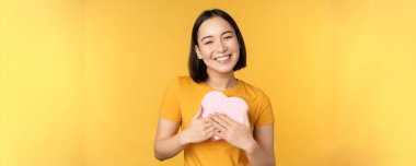 Romantizm ve sevgililer günü. Büyük bir kalp kartı tutan, gülümseyen, sarı arka planda duran mutlu, güzel Asyalı kadın.